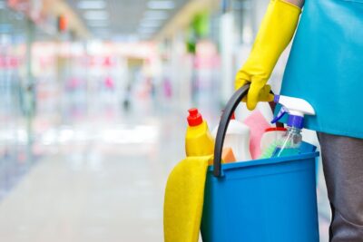 Mujer con guantes de goma azules sosteniendo un balde con productos de limpieza para el hogar.